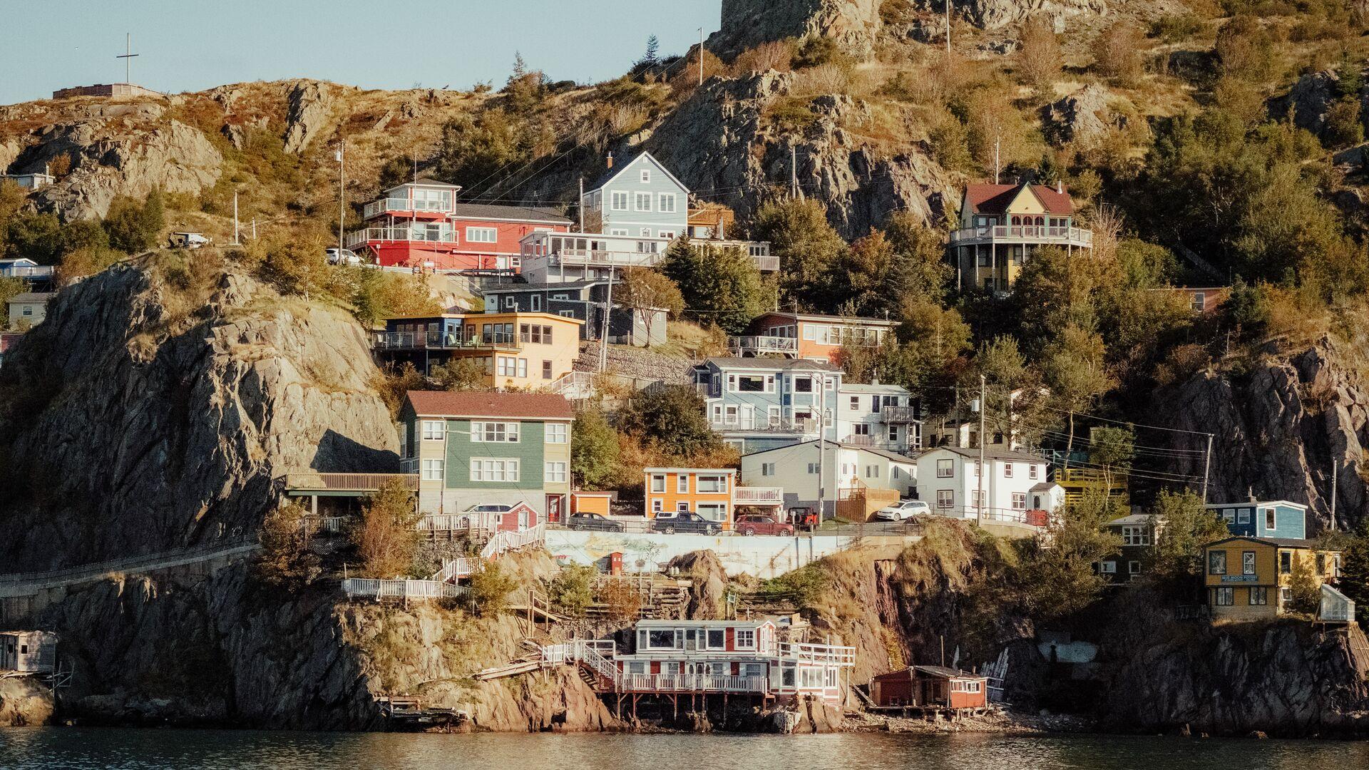 Colorful houses sit on a cliff beside the ocean. Between the buildings there are trees and long grass.