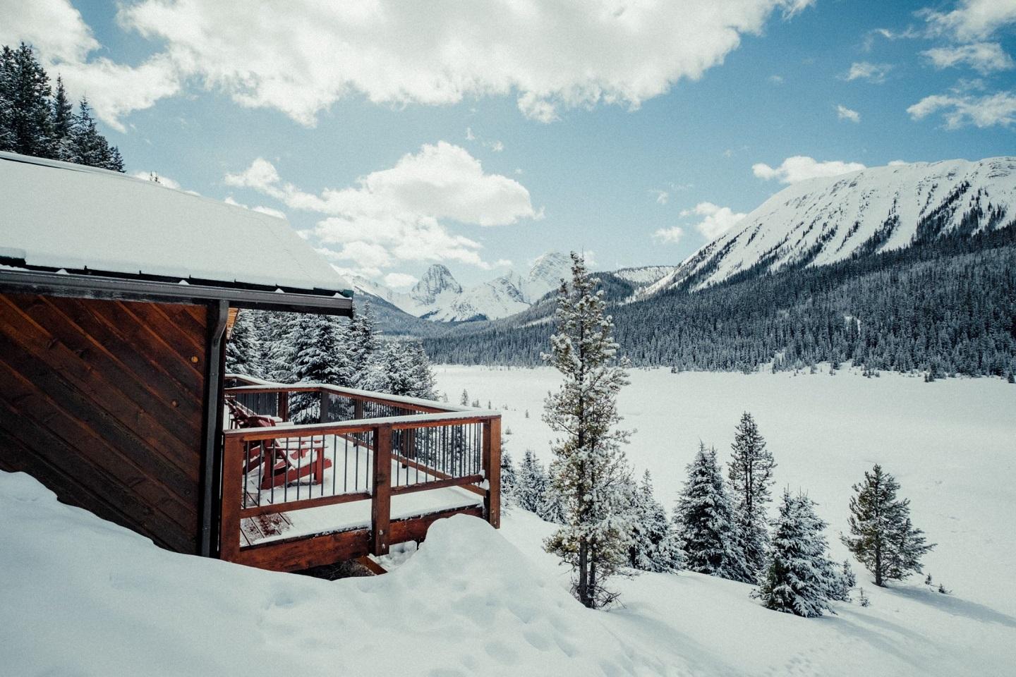 A wood cabin covered in snow overlooking a frozen over lake on a clear day.