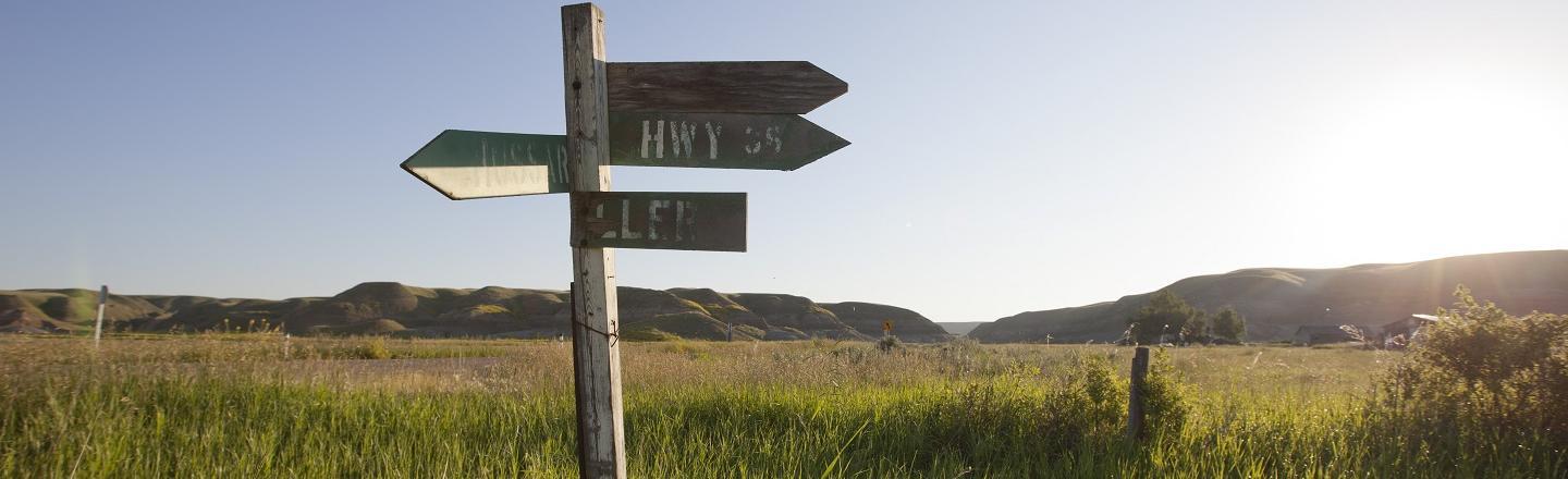 A rustic old highway sign in front of a sprawling field. There are multiple small signs pointing in different directions with rubbed off labels.