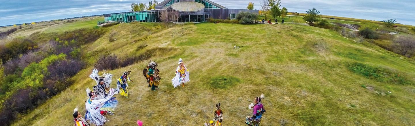A group of people dressed in indigenous regalia dancing in a field outside of a building