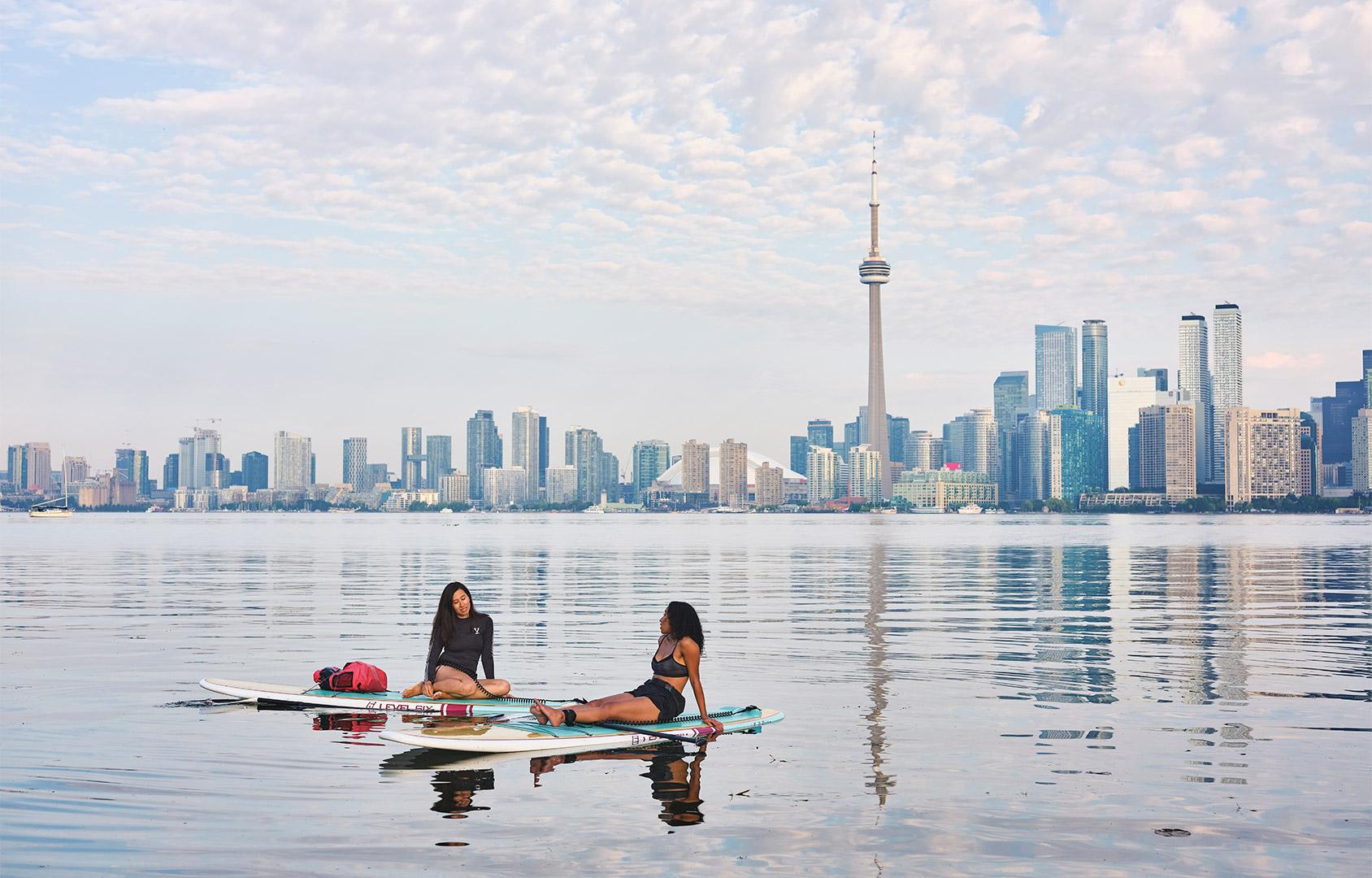 Two people sit on paddleboards on calm water with the Toronto skyline and CN Tower in the background under a partly cloudy sky.