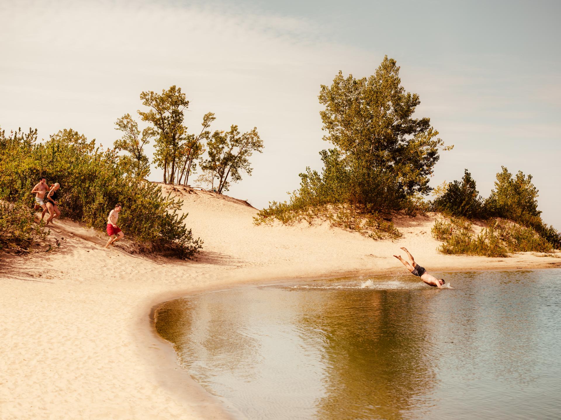 Three people dressed in swimwear run down a sand dune towards a lake. A fourth person ahead of them is diving into the water, and the sand is surrounded by green trees and bushes.